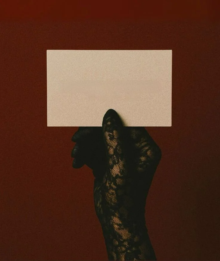 A hand wearing a black lace glove holding a blank white card against a dark red background.