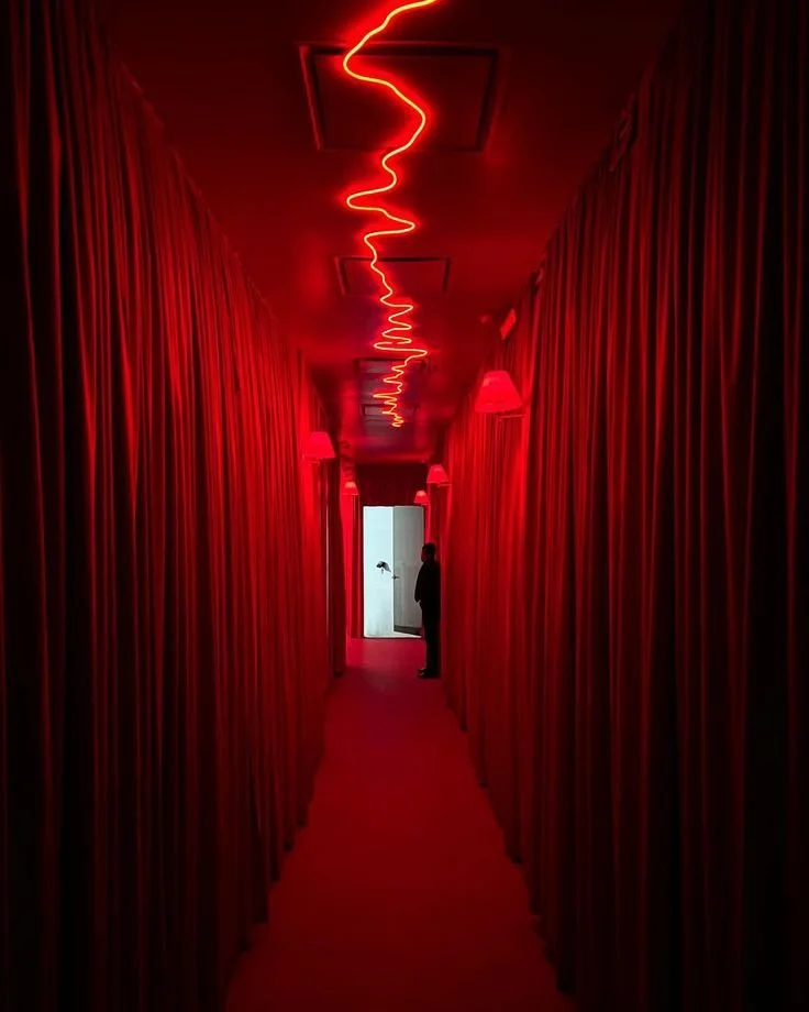 Dimly lit red hallway with curtains on both sides, a person standing at the end near a door with white light shining through.