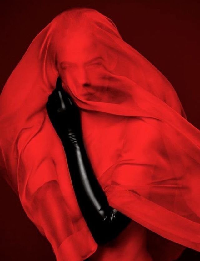 Person wrapped in a red translucent fabric with black gloves, against a dark background.