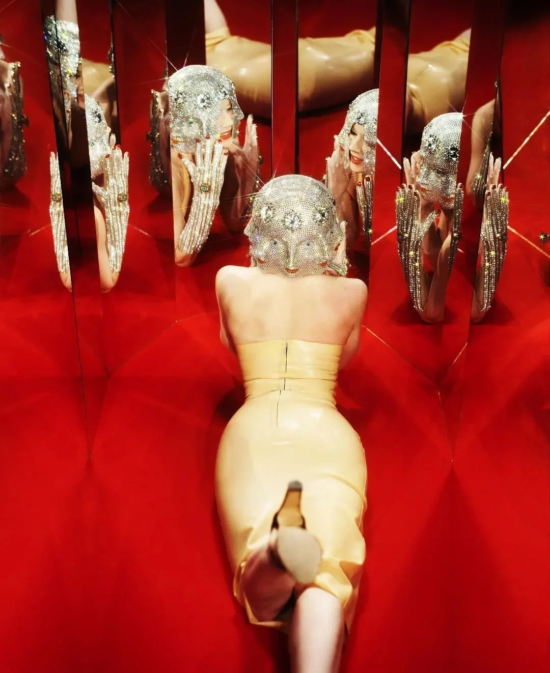 A woman in a short yellow dress with a sparkling rhinestone mask is kneeling on a red surface with multiple mirrors reflecting her and her mask, creating a kaleidoscopic effect.