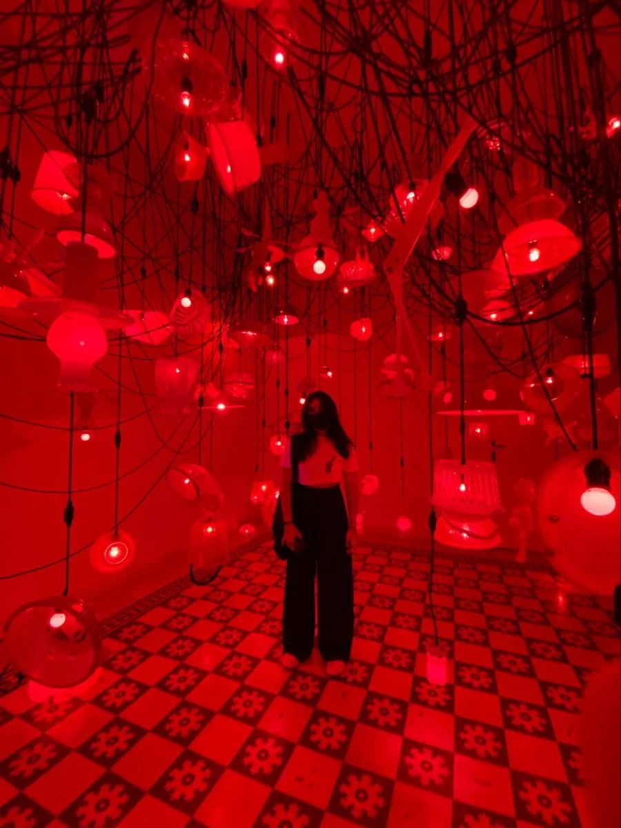 Woman standing in a red-lit room with hanging red lamps and tangled black wires, checkered floor.