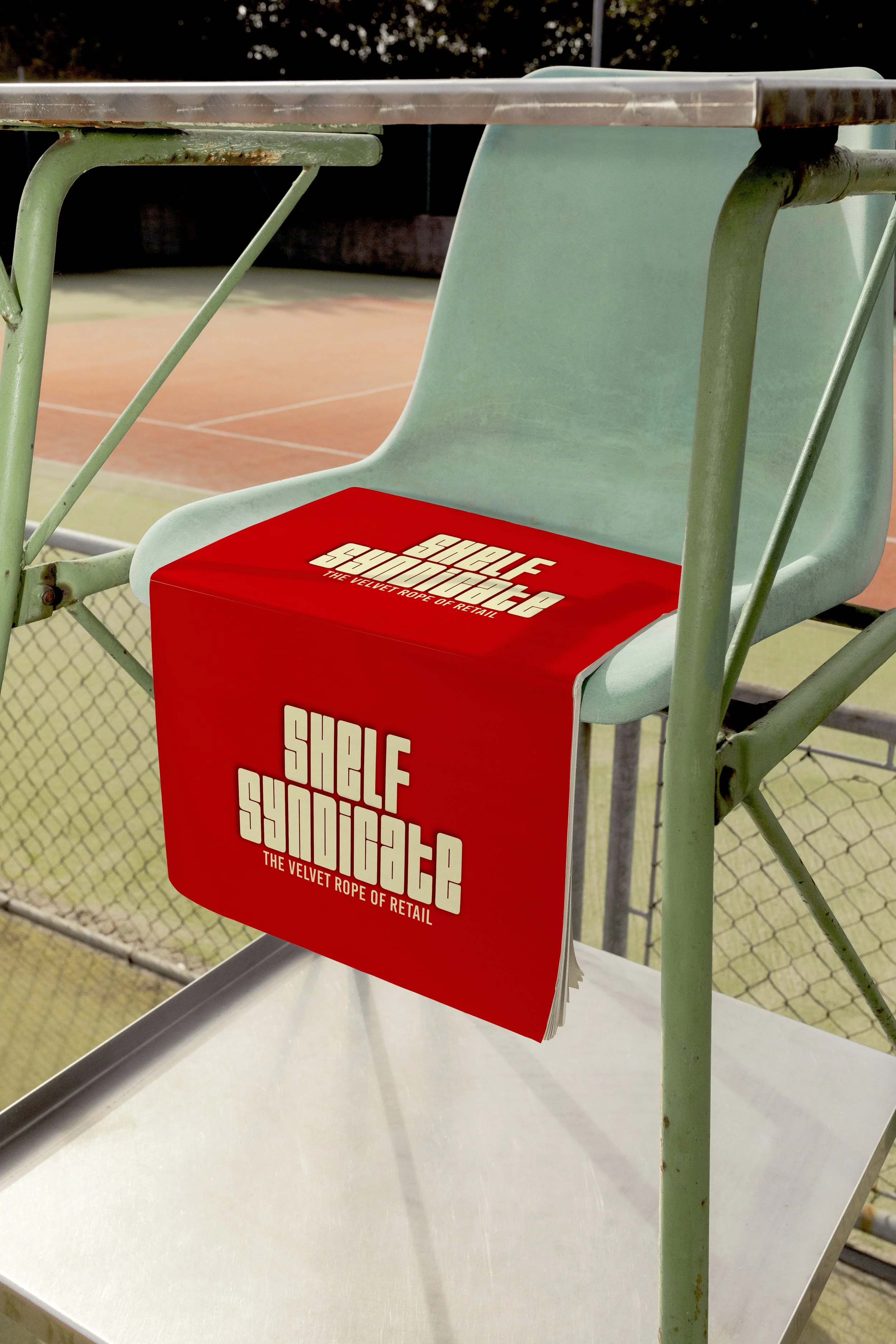 A red cardboard box labeled "SHELF SYNDICATE: THE VELVET ROPE OF RETAIL" hanging on a green metal chair near a tennis court.