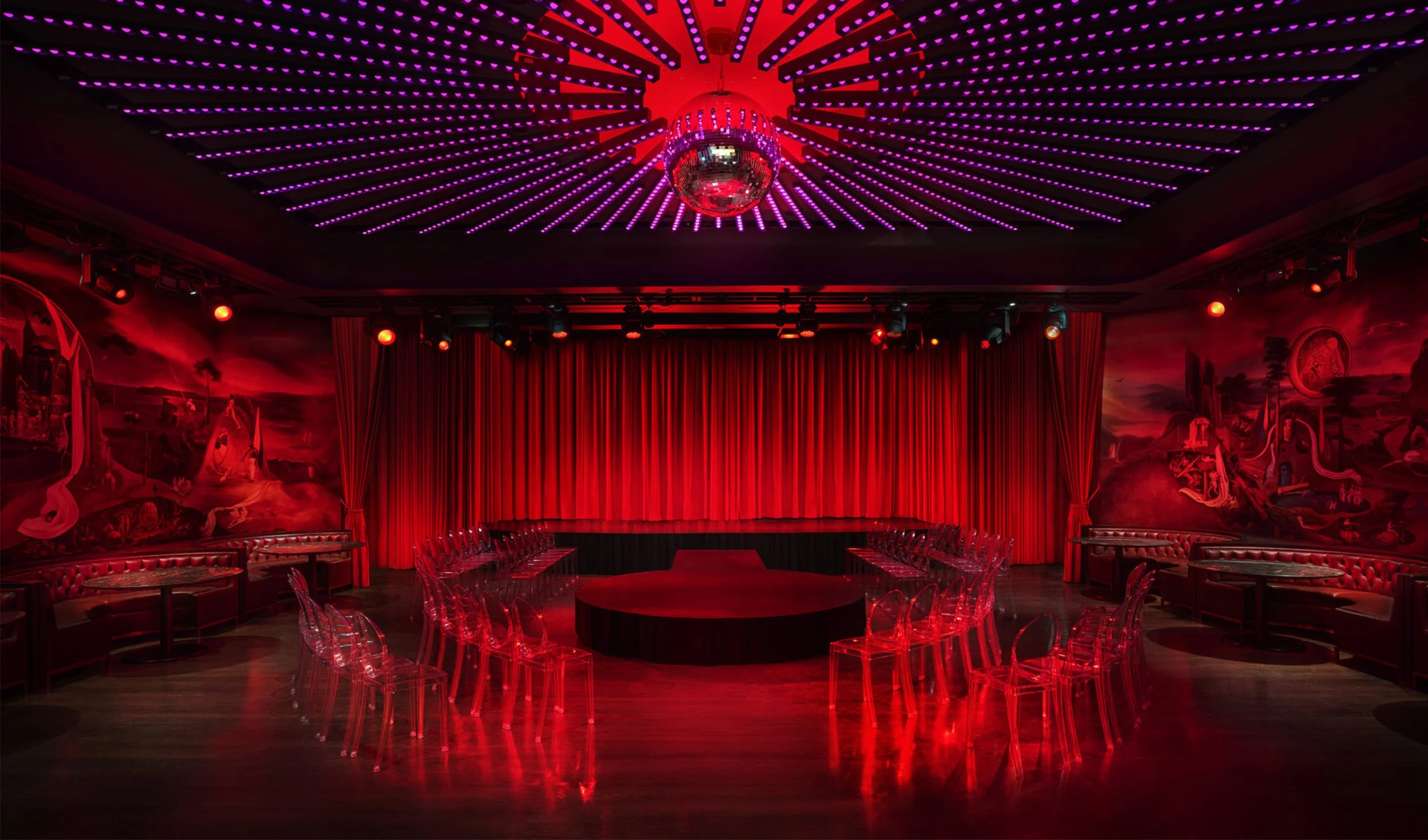 A dimly lit entertainment or performance room with a red stage curtain, red ambient lighting, transparent chairs arranged around a circular table, and a ceiling with pink LED lights and a disco ball.