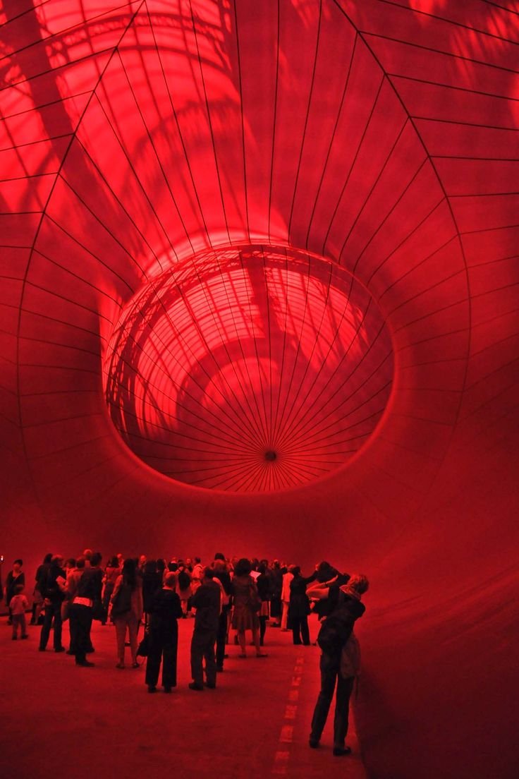 People standing inside a large, red-lit, spiral-shaped installation or tunnel with geometric patterns and a reflective surface.