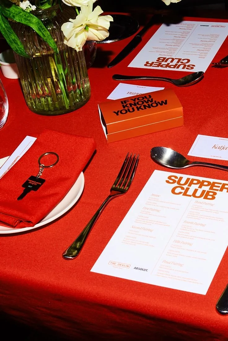Table setting with a red tablecloth, a vase with white flowers, a printed menu titled 'Supper Club,' a small orange card that says 'If you know, you know,' a keychain, and utensils including a fork, spoon, and knife.