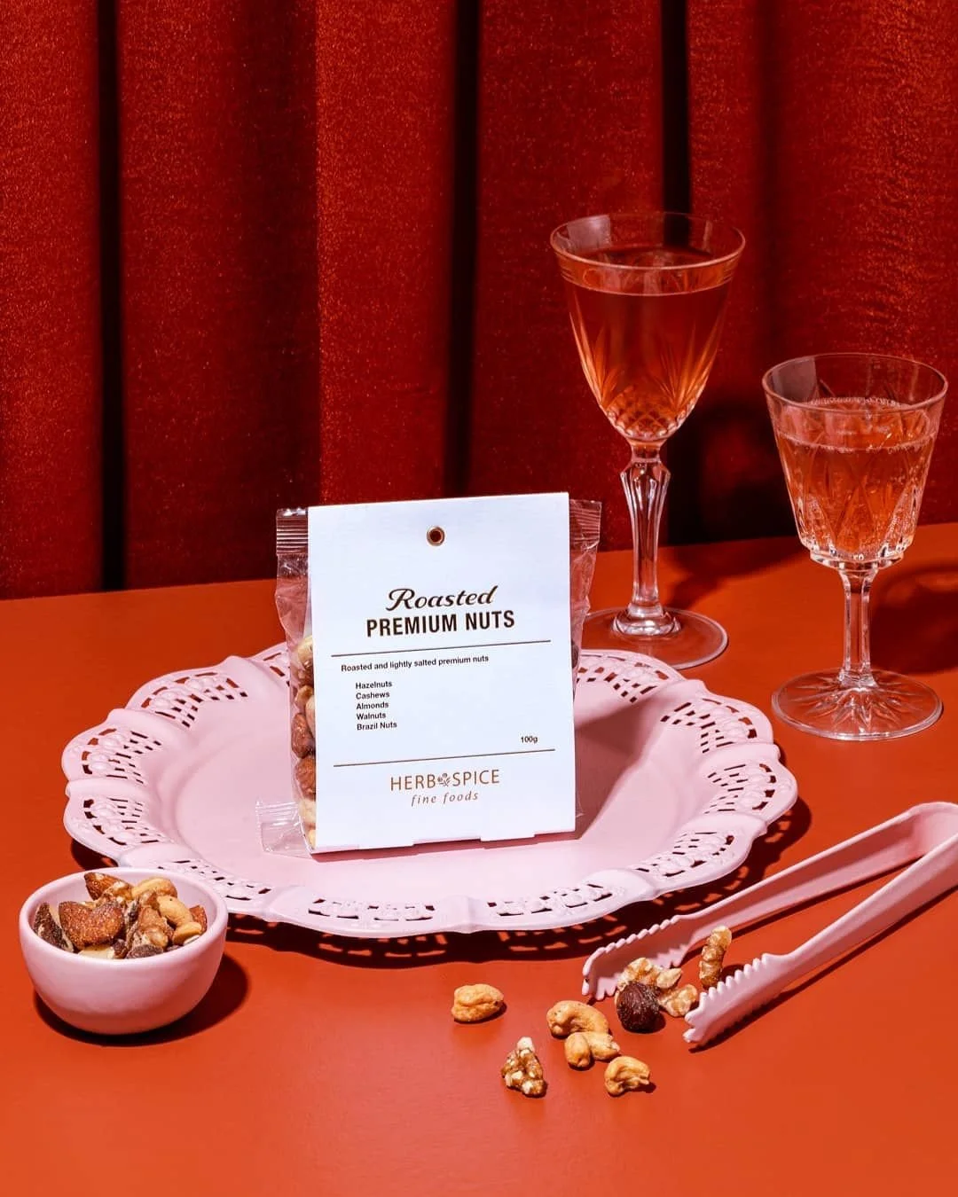 Package of roasted premium nuts on a white lace tray, with a small bowl of mixed nuts nearby, two glasses of pinkish drink, and pink tongs on a reddish-brown surface with a red velvet background.