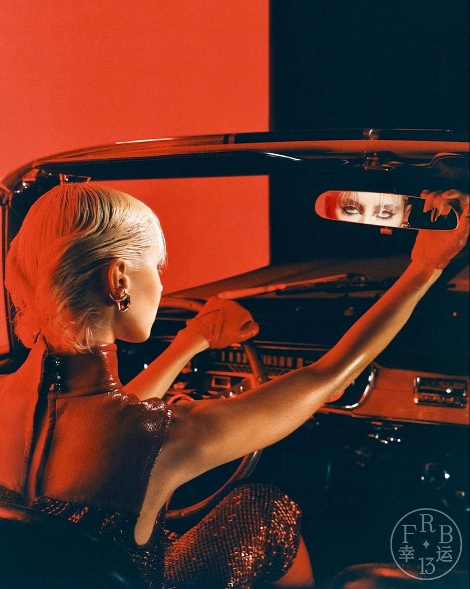A woman with platinum blonde hair and gold hoop earrings is inside a car, adjusting the rearview mirror. The mirror reflects her eyes. The scene is lit with red and orange lighting.