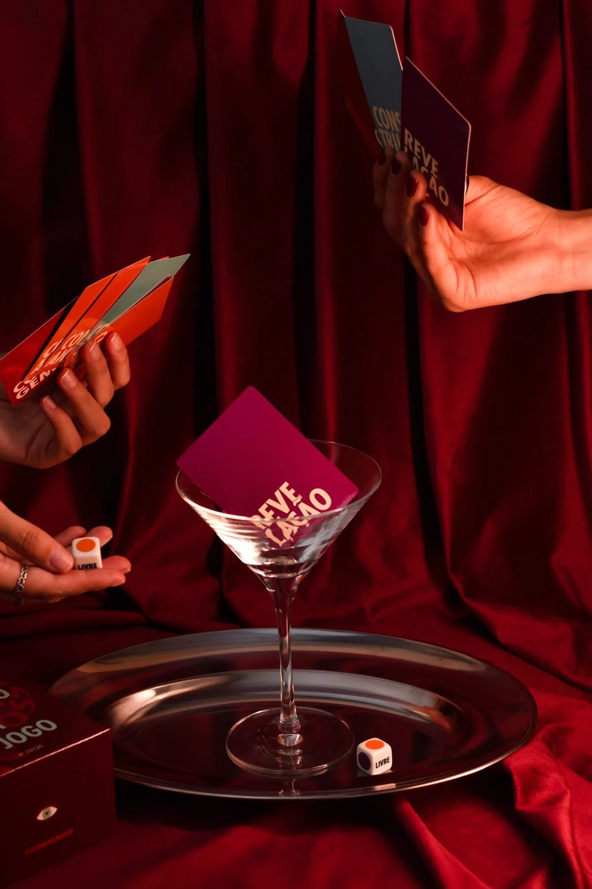 Hands holding colorful cards and dice displaying the words 'LIVRE' and 'CONSEIL' against a red velvet background with a martini glass and a silver tray.