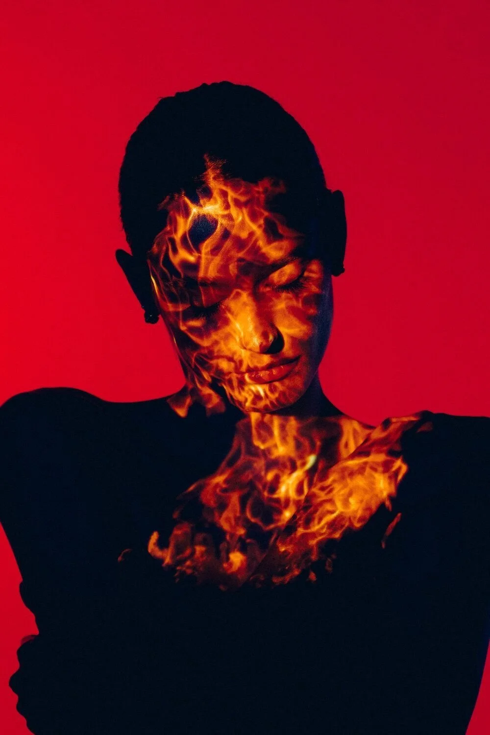 A person with short hair and closed eyes, with flaming fire superimposed on their face and neck against a red background.