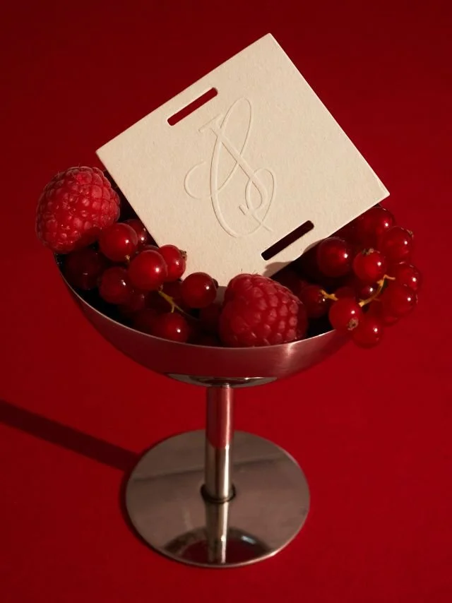 A metal bowl filled with raspberries and red currants, placed on a red surface. A white card with an embossed cursive letter "A" is resting inside the bowl.