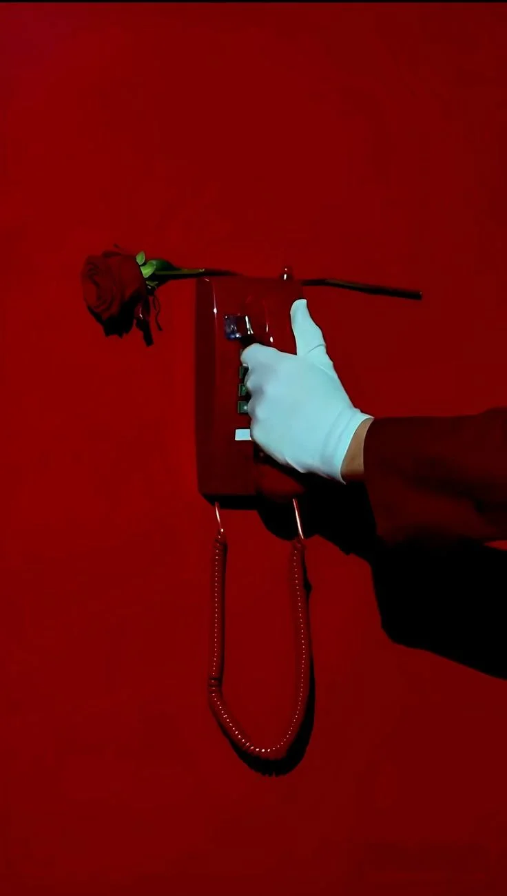 A hand wearing a blue glove holding an old-fashioned red telephone with a red rose placed on top of it, all against a red background.