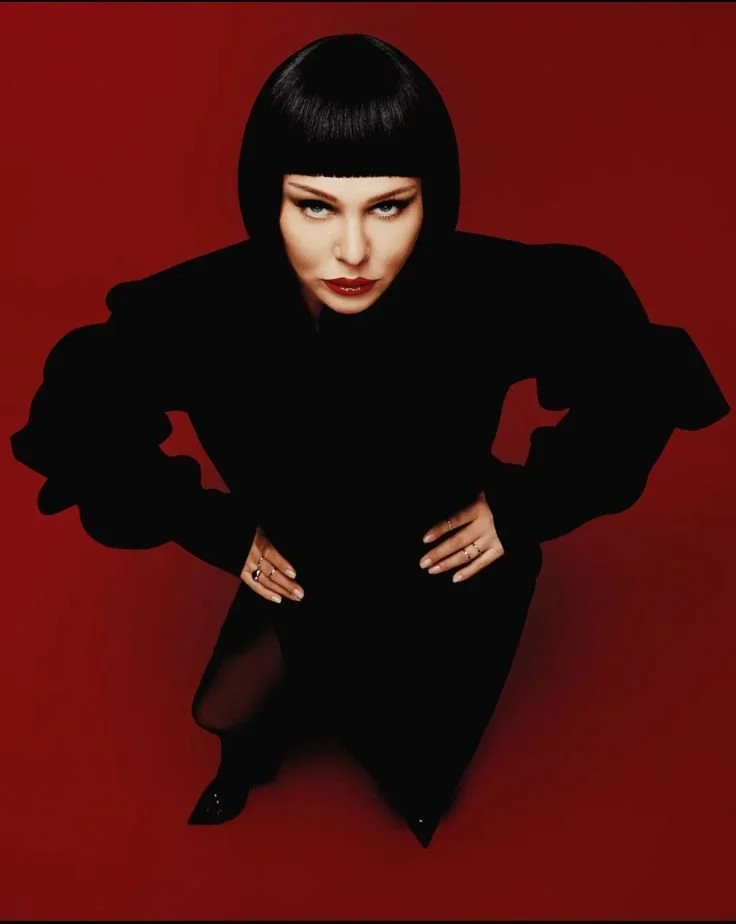 A woman with a short black bob haircut and bold makeup posing against a red background, wearing a black outfit and looking directly at the camera.