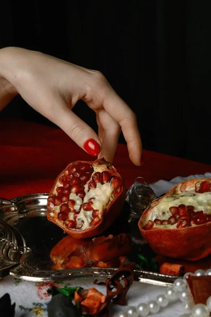 A hand with red-painted nails holding a halved pomegranate over a silver tray with more pomegranate halves on a decorated table.