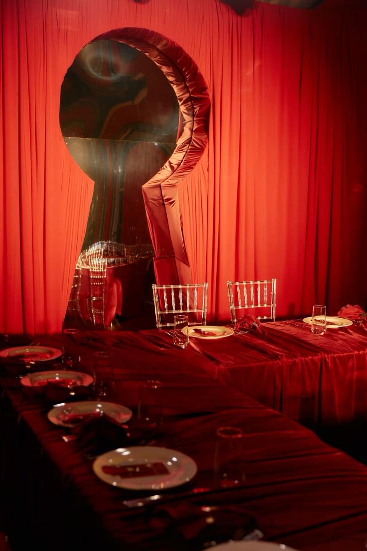 Table set with plates, glasses, and utensils in front of a red curtain with a large, artistic mirror or sculpture resembling a question mark.