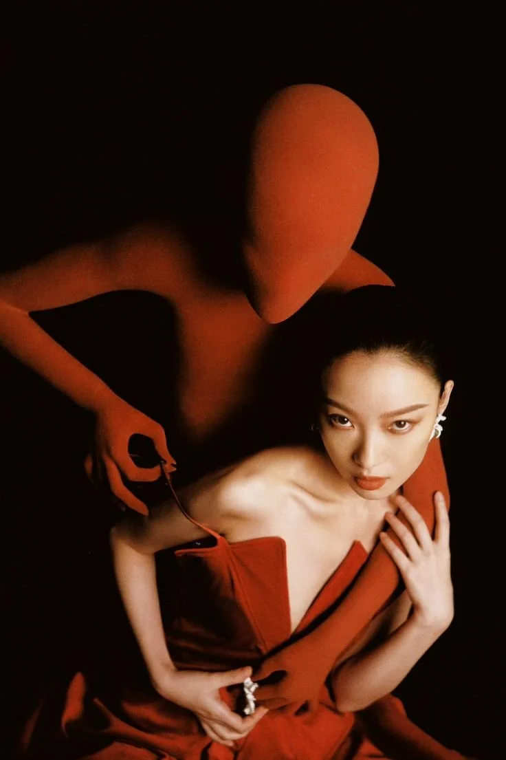 A woman in a red dress, sitting with her hand on her chest, looking directly at the camera, with an orange humanoid figure standing behind her, head tilted, on a black background.