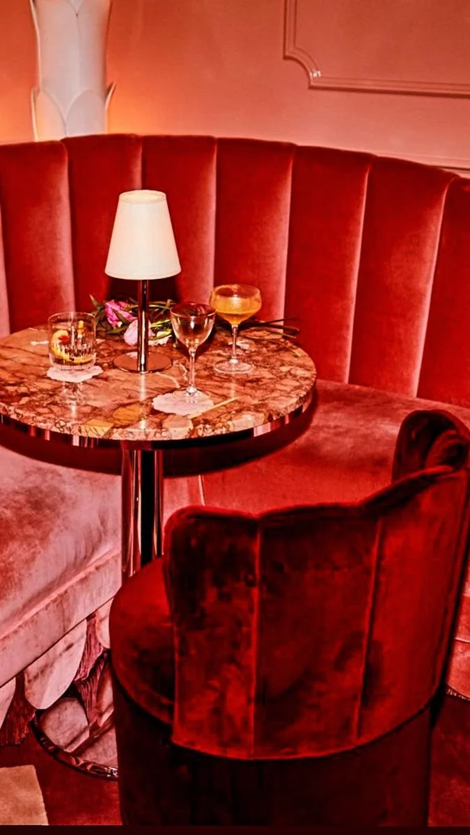 A cozy dining area with a round marble table, a red velvet booth, and a matching red velvet chair. On the table, there is a small lamp, four glasses, and some flowers with two long stems.