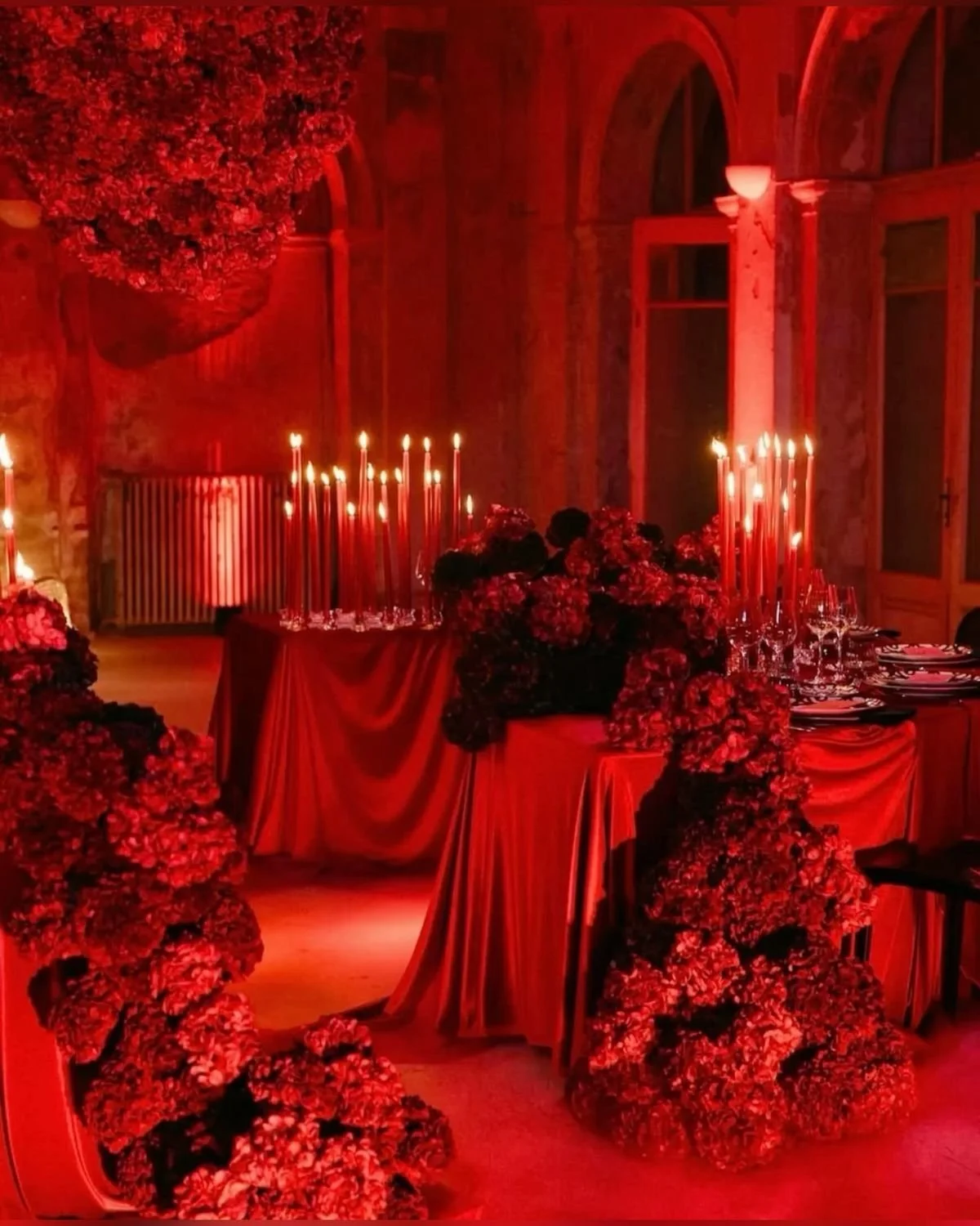 A romantic dining setup in a dimly lit room with red lighting. Features a table with floral arrangements, tall red candles, and elegant tableware, all decorated with red and dark flowers, creating an intimate atmosphere.