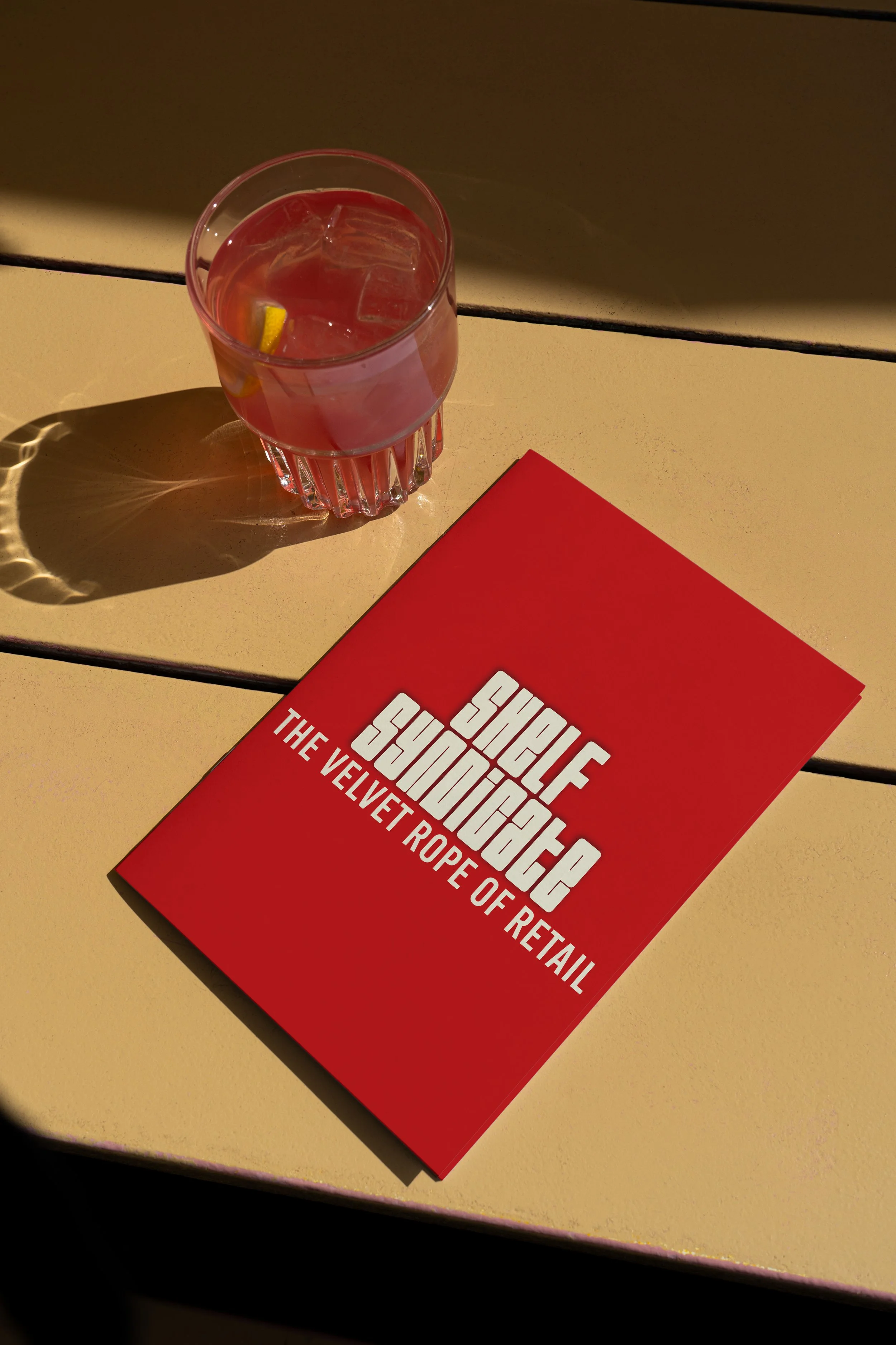 A red book titled "Super Sumo Summer: The Velvet Rope of Retail" lying on a beige tabletop with a glass of pink drink with ice and lemon slices, casting a shadow.
