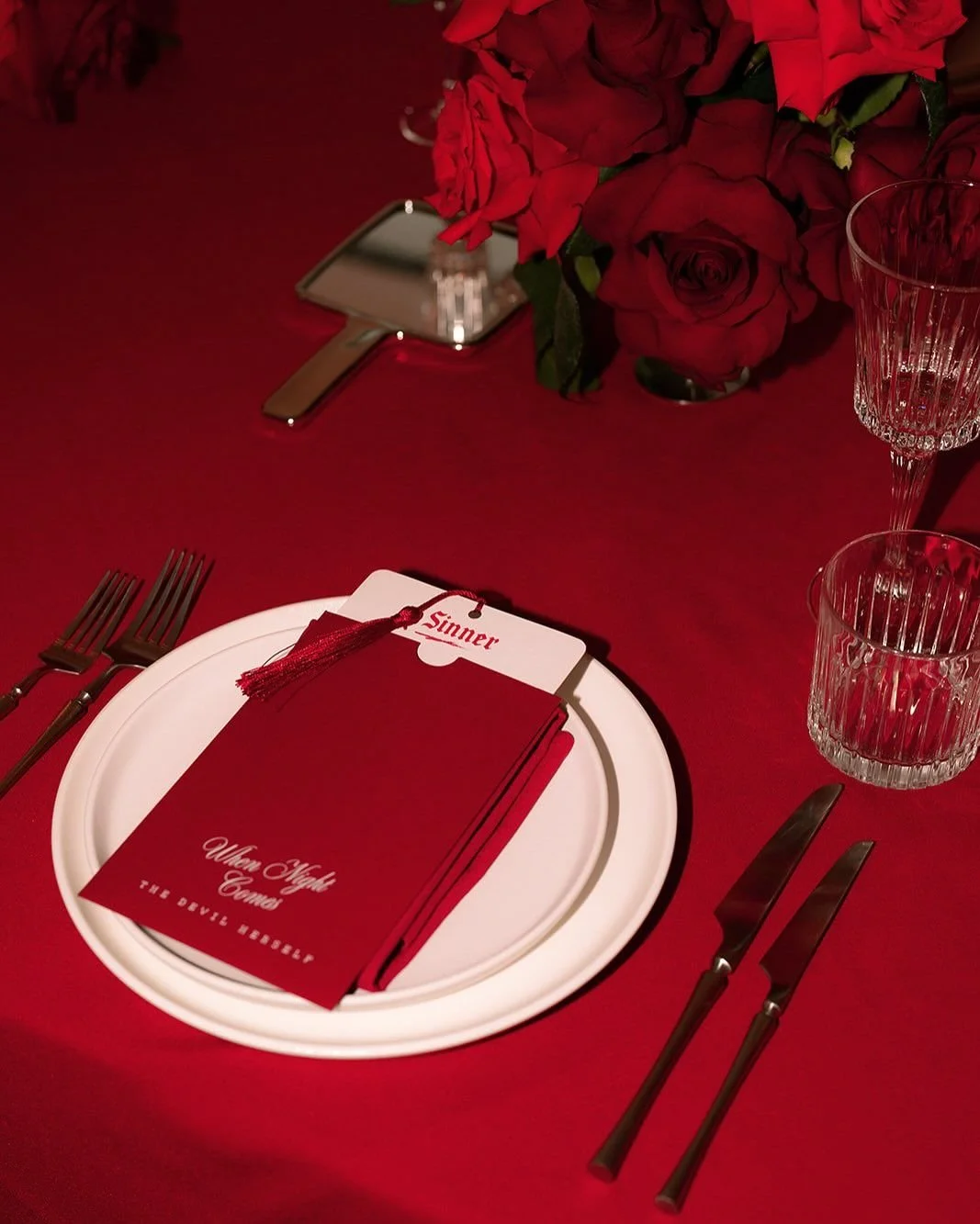 A red-themed table setting with a white plate, red napkin, and a red accordian with 'When Wolf Comes' and 'The Devil Wears' written on it, a poker card with 'Sinner,' a bunch of red roses, crystal glasses, and a small mirror.