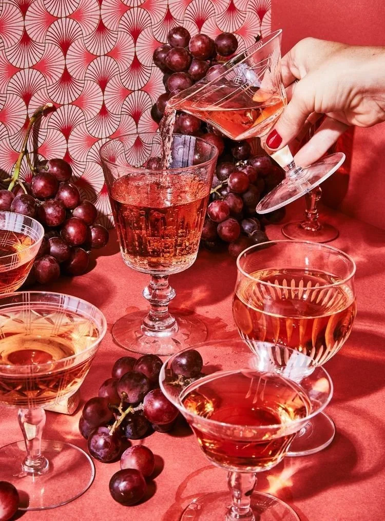 A hand pouring red wine or another red beverage from a small glass into a larger glass, surrounded by grape clusters and additional glasses of the same drink on a red surface with a red patterned background.