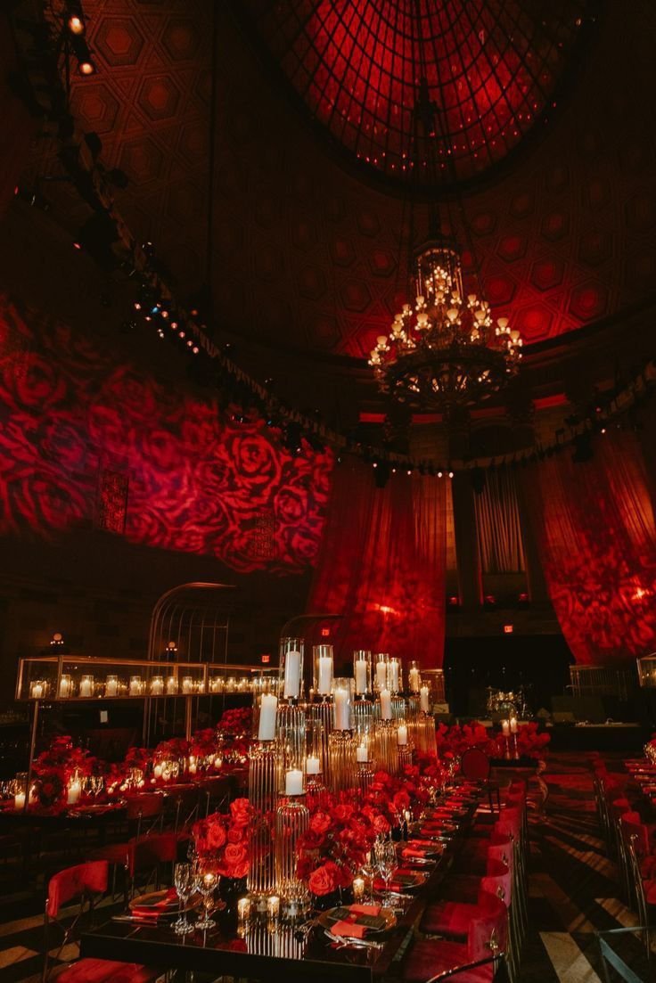 Elegant dining hall decorated for a formal event with red roses, candle centerpieces, and draped red curtains. The ceiling features a large chandelier and patterned red lighting projections.