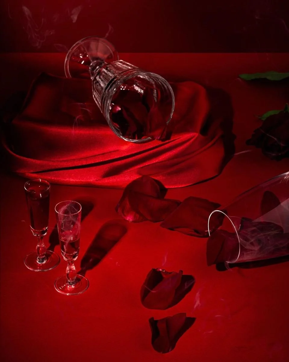 A photo features an overturned wine glass on a red velvet cloth with rose petals and two empty champagne glasses nearby, all set on a red surface with a dark background.