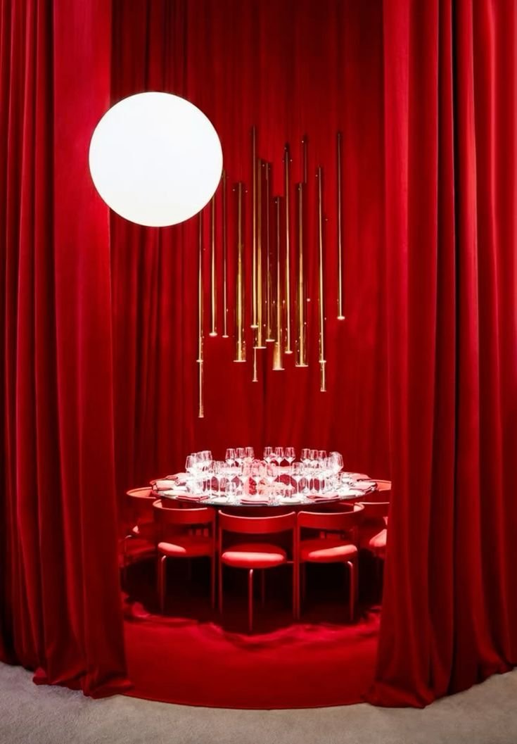 Elegant red-curtained dining area with a round table set with glasses and surrounded by red chairs, illuminated by a large white spherical light and modern hanging gold pendant lights.