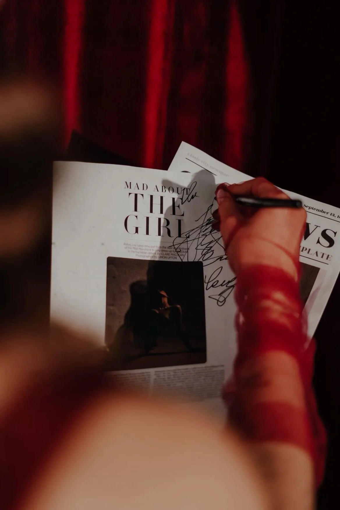 Person signing a newspaper with the headline 'Mad About the Girl' while holding it in their left hand, with a dark background and red curtains.