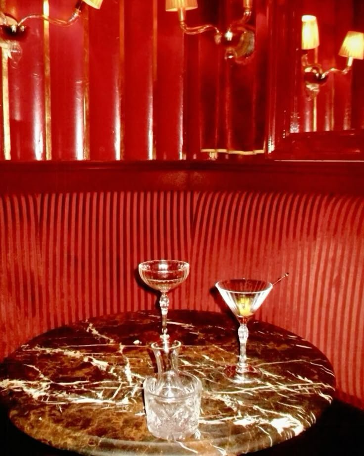 Two empty cocktail glasses and one empty short glass on a dark marble table in a dimly lit, red-themed lounge or bar setting.