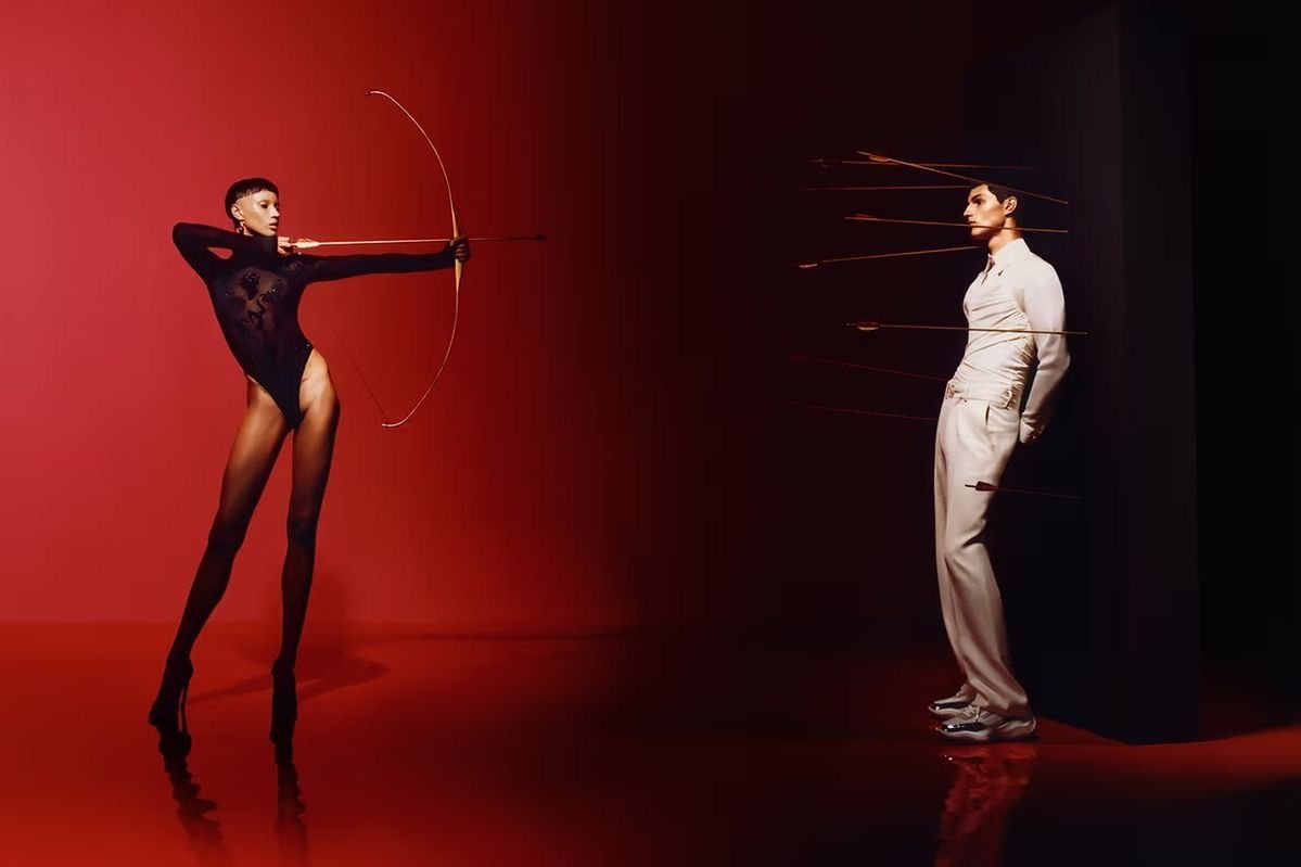 A woman in black lingerie aiming a bow and arrow at a man in a white suit who has arrows piercing his side.