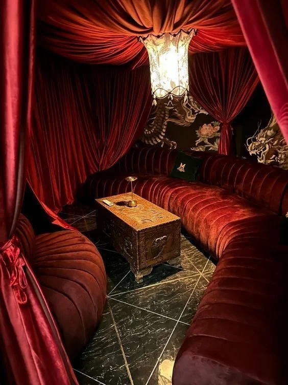 Luxurious red velvet curtains and seating in a dimly lit room with a dragon mural on the wall and a small wooden table in the center.