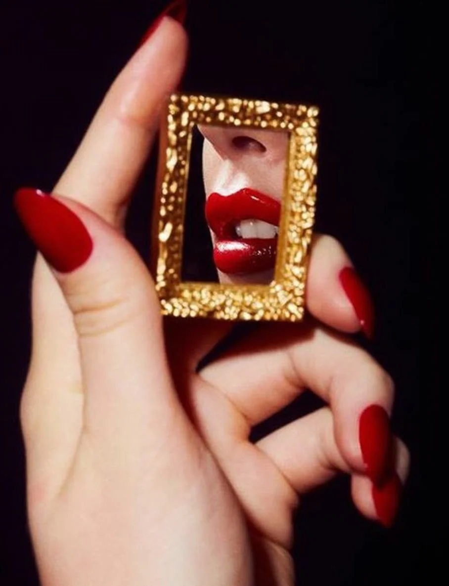 A person holding a small gold glittery picture frame with red nail polish, showing a reflection of lips with bold red lipstick and partially visible nose on a black background.