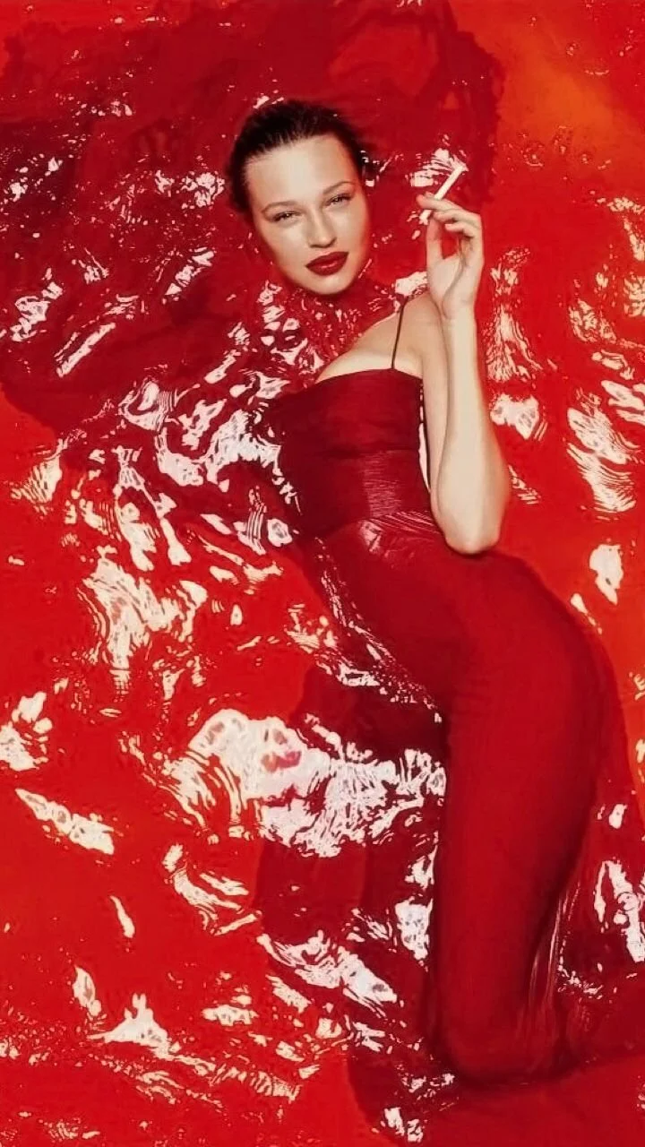 A woman lies in red water, wearing a matching red dress and lipstick, holding a cigarette.