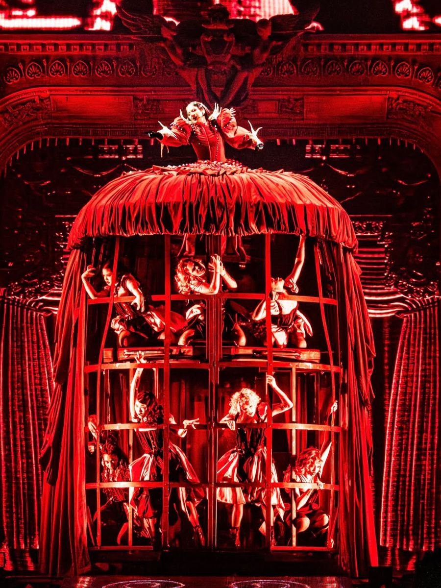 A theatrical performance featuring multiple performers dressed in elaborate costumes, inside a large cage with red velvet curtains. The scene is set on a richly decorated stage with ornate architectural details.