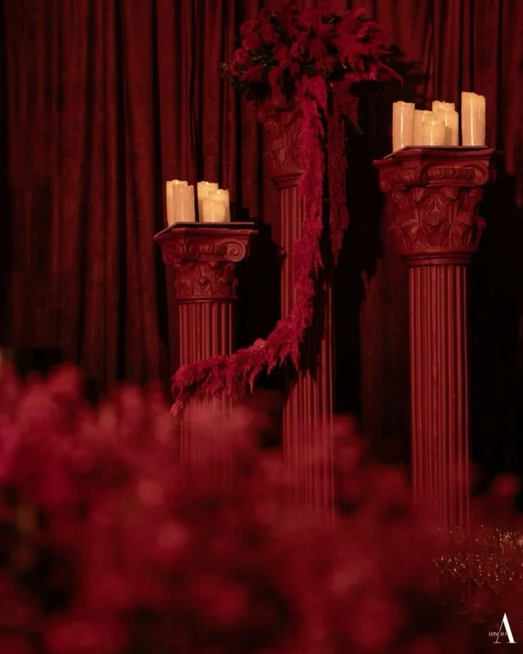 Decorative scene with three red classical columns, draped with red feathered garland, candle arrangements, and a bouquet of red flowers against red velvet curtains.