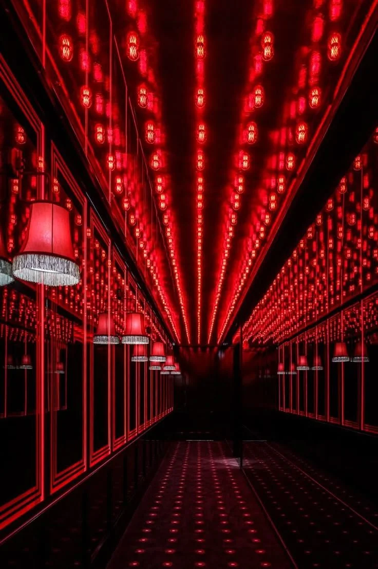 A hallway illuminated with red LED lights on the ceiling, reflecting in mirrored walls, creating a tunnel effect with a person walking in the distance.