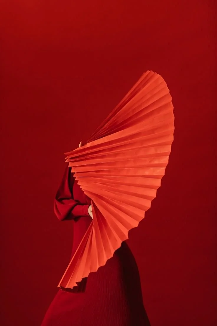 Person dressed in red holding a large red folding fan, standing against a red background.