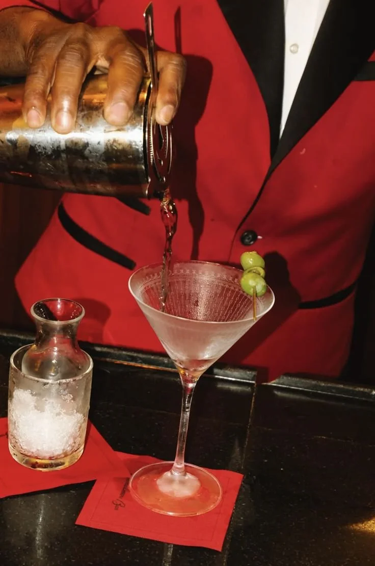 Bartender pouring a cocktail into a martini glass garnished with green olives at a bar.