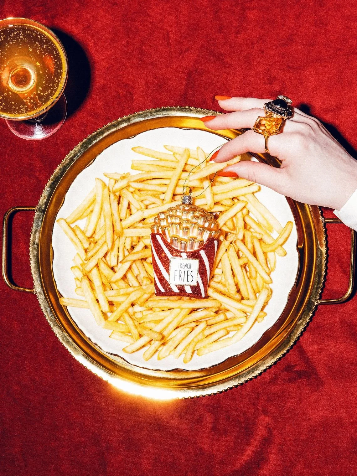 A tray of French fries with a small Eiffel Tower ornament on top, placed on a white paper lining, with a hand wearing rings reaching for the fries, and a glass of champagne nearby on a red table.