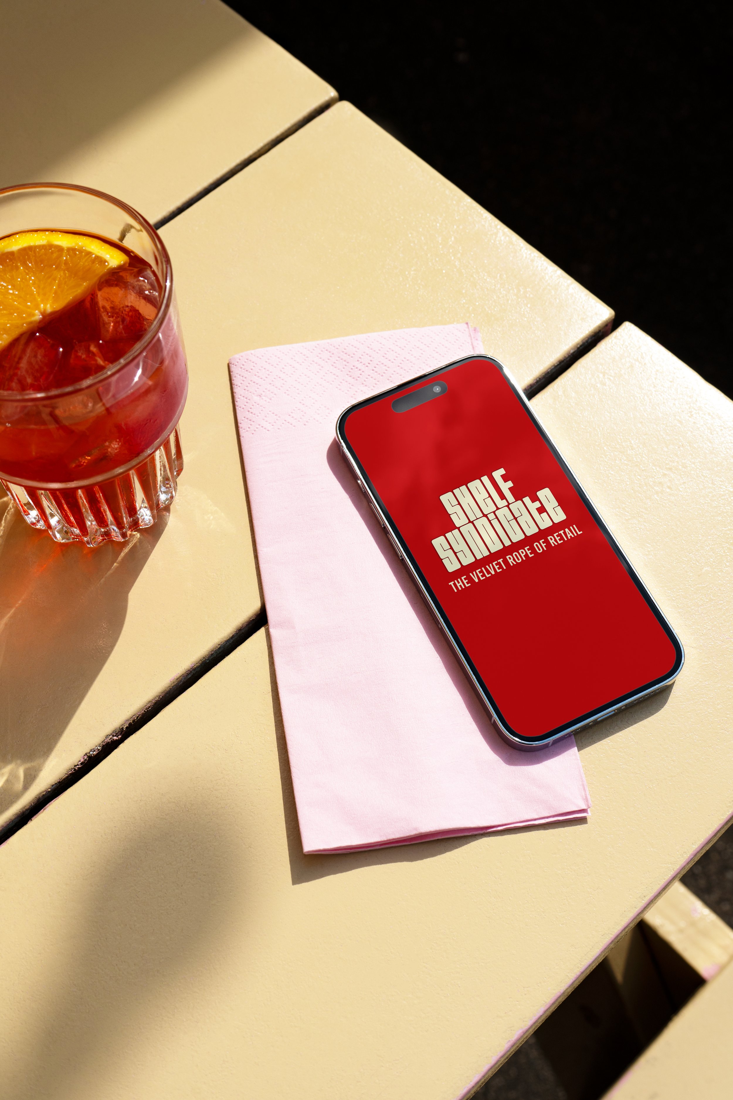A smartphone displaying the logo and tagline of Single Squadron resting on a pink cloth napkin on a yellow outdoor table, with a glass of iced tea with lemon and ice cubes nearby.