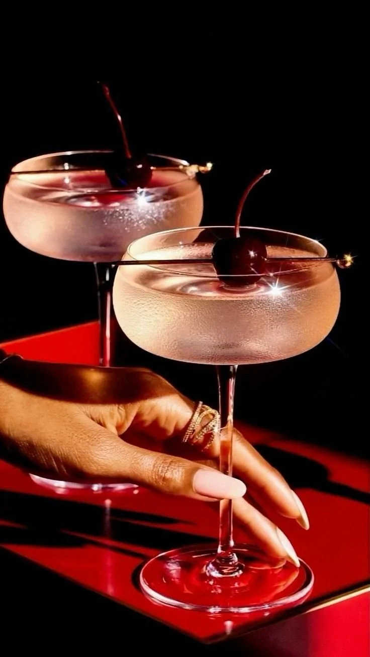 Two cocktail glasses with cherries, being held by a person with rings on fingers, against a black background.