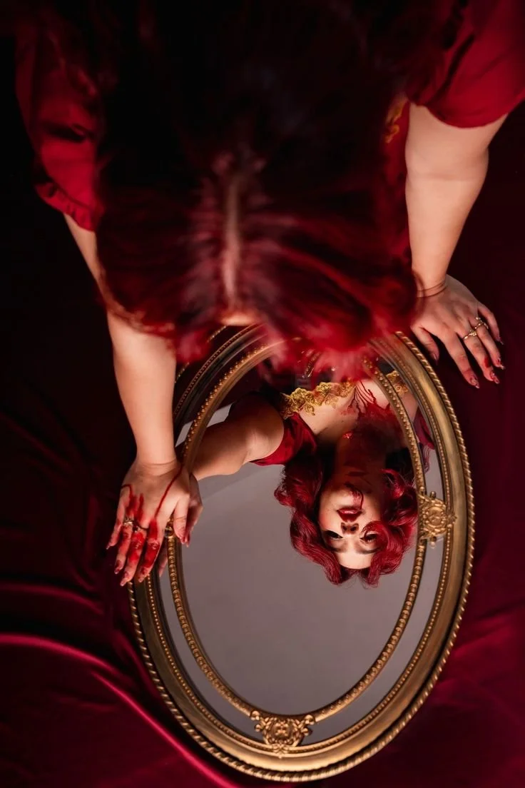 A woman with red hair looking upwards at her reflection in an ornate oval mirror, surrounded by red drapery.