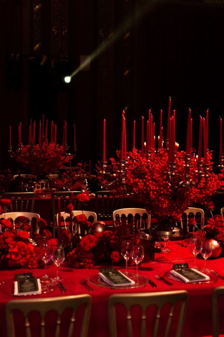 A darkly lit banquet table decorated with tall red candles, red floral arrangements, and elegant glassware, set for a formal event.