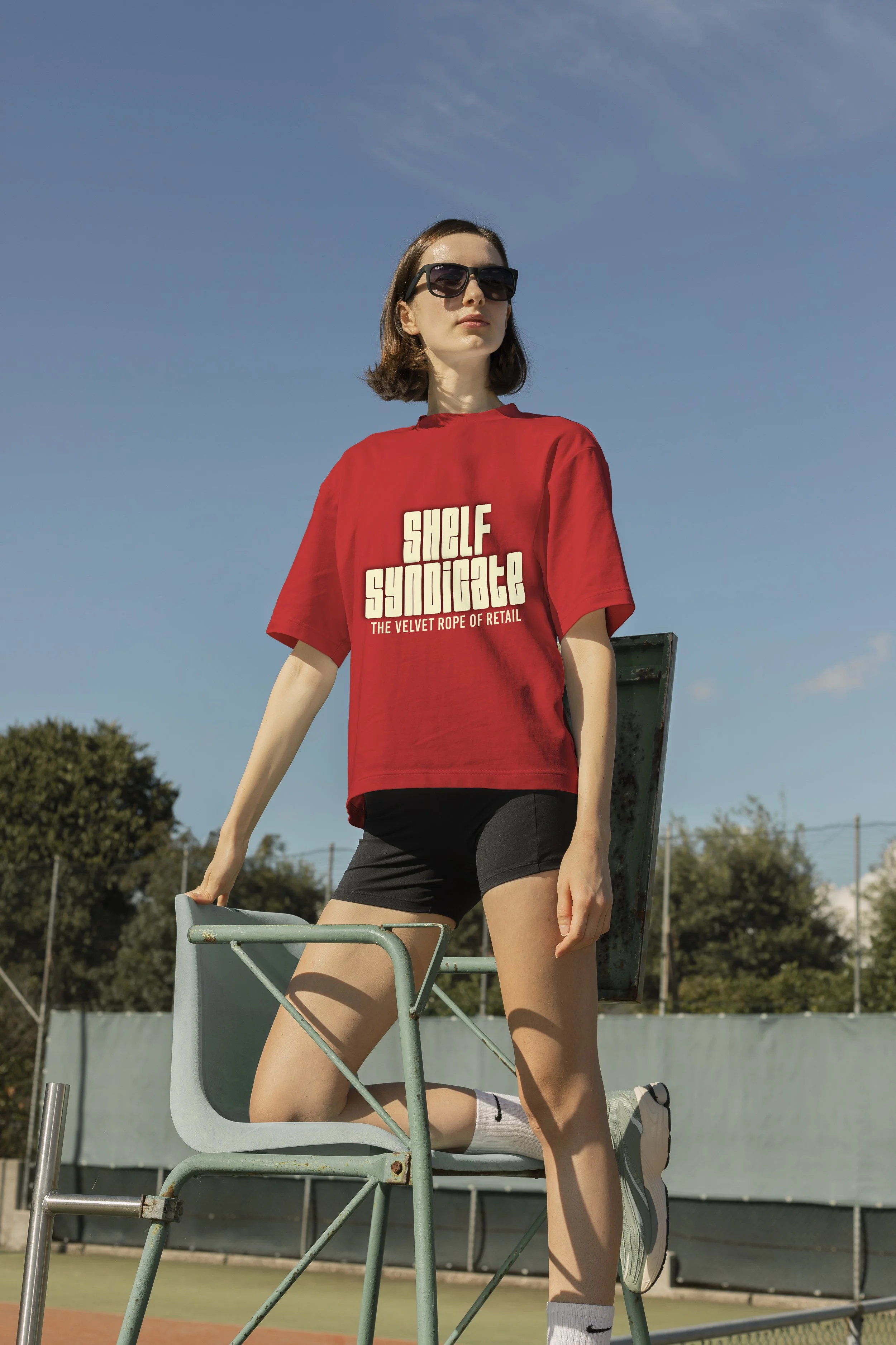 A young woman with sunglasses and a red t-shirt standing on a tennis umpire chair outdoors on a sunny day.