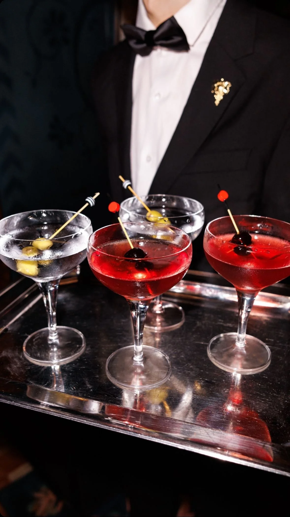 Three cocktail glasses with garnished cherries and olives, served on a silver tray, in front of a person dressed in formal attire with a bow tie.
