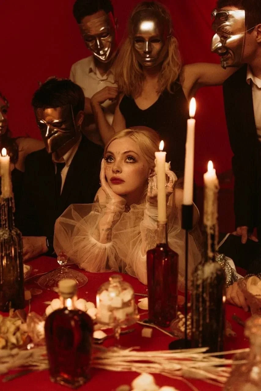 A woman with blonde hair gazes upward with a dreamy expression, surrounded by people wearing metallic masks at a dimly lit, elegant dinner party with candles and ornate decorations.