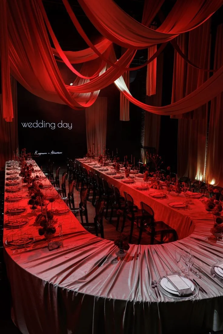 Elegant wedding reception setup with a curved table draped in white fabric, adorned with floral centerpieces, set for a formal dinner, and a backdrop with the words 'wedding day' illuminated.