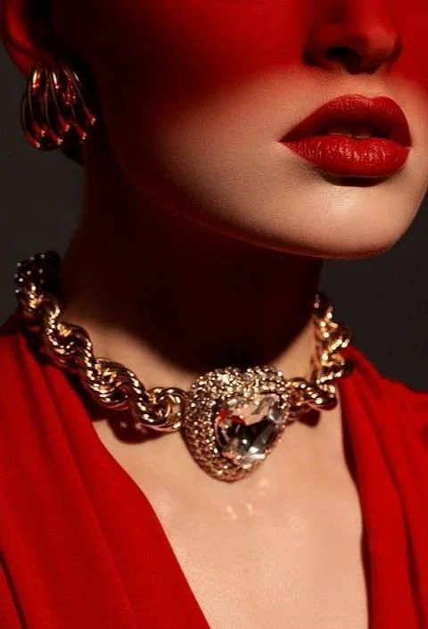 Close-up of a woman wearing a bold red lipstick, a chunky gold chain necklace with a large ornate heart-shaped pendant, and matching gold earrings. She is dressed in a red outfit against a dark background.