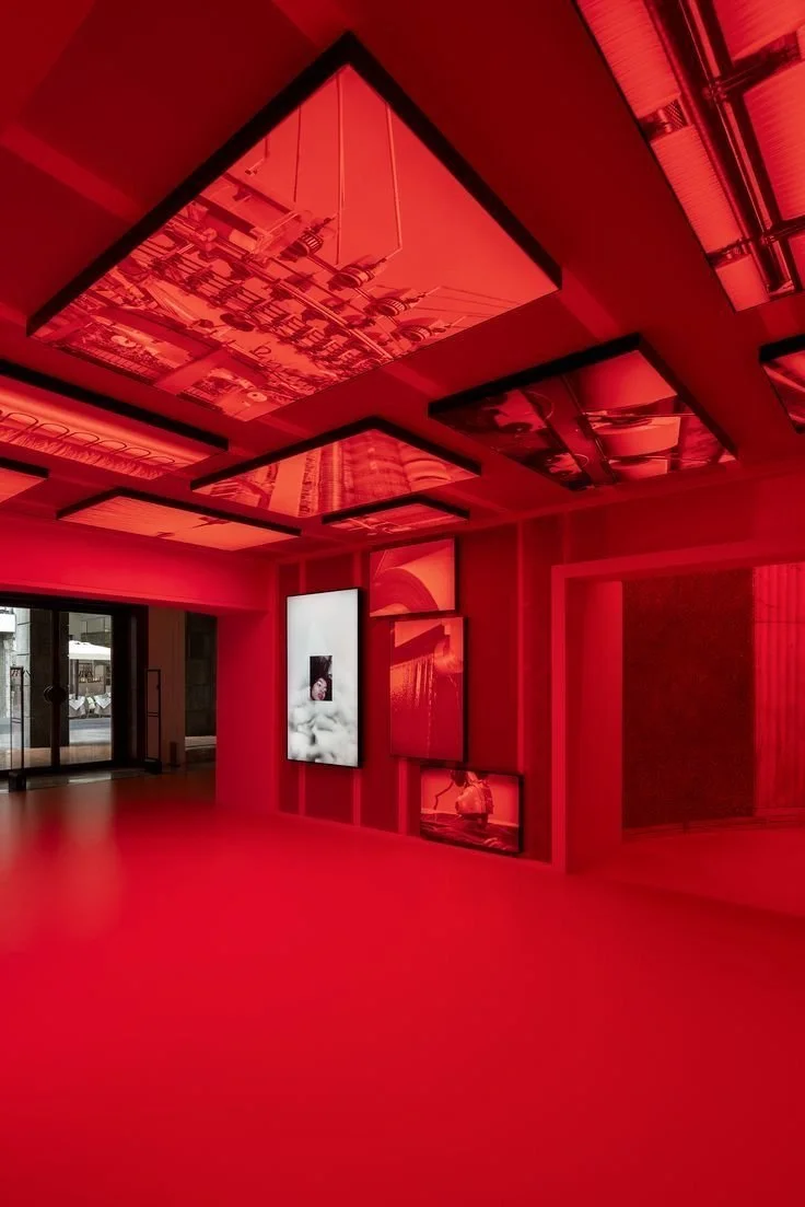 Modern art gallery with red lighting and framed artwork on the walls, glossy ceiling panels reflecting images, glass doors leading outside.