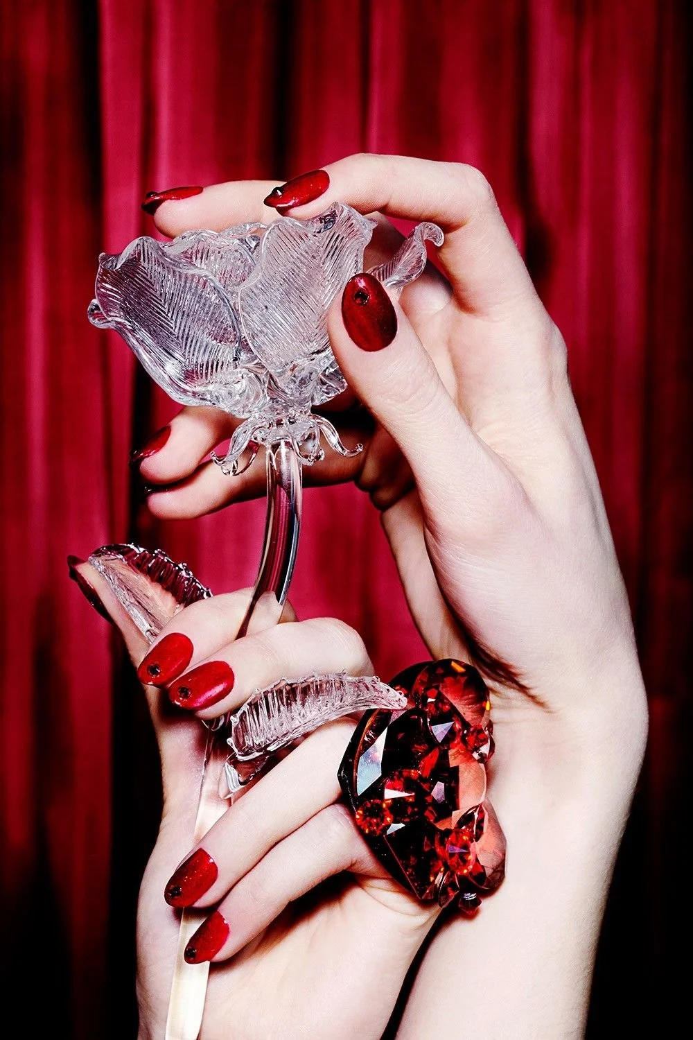 A person's hand holding a decorative, art glass flower with red painted nails and a large red heart-shaped ring against a red curtain background.